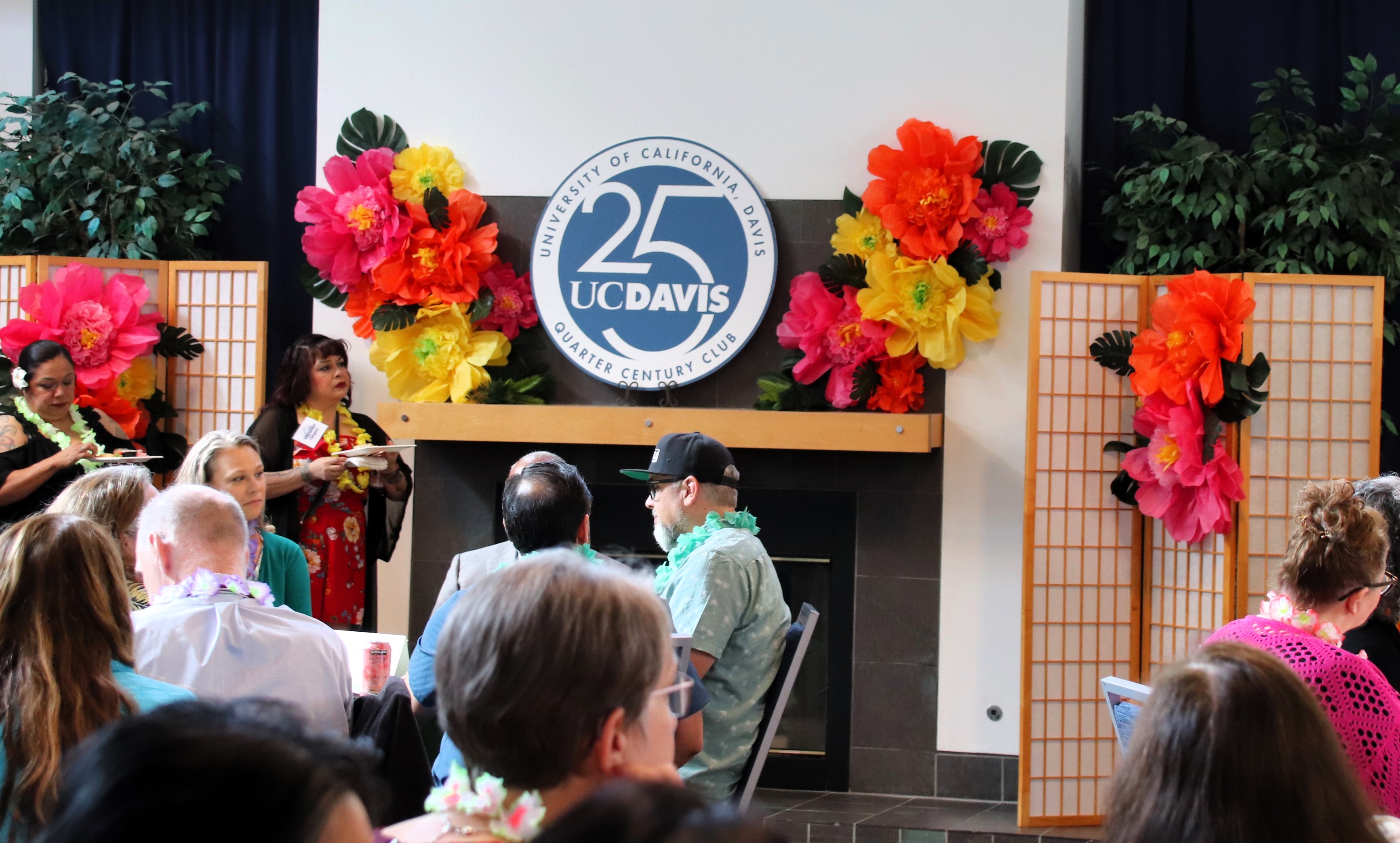 Orange and yellow flowers and a white and blue round sign that reads "Quarter Century Club" with a large 25 in the center adorn the mantle at the front of the room.