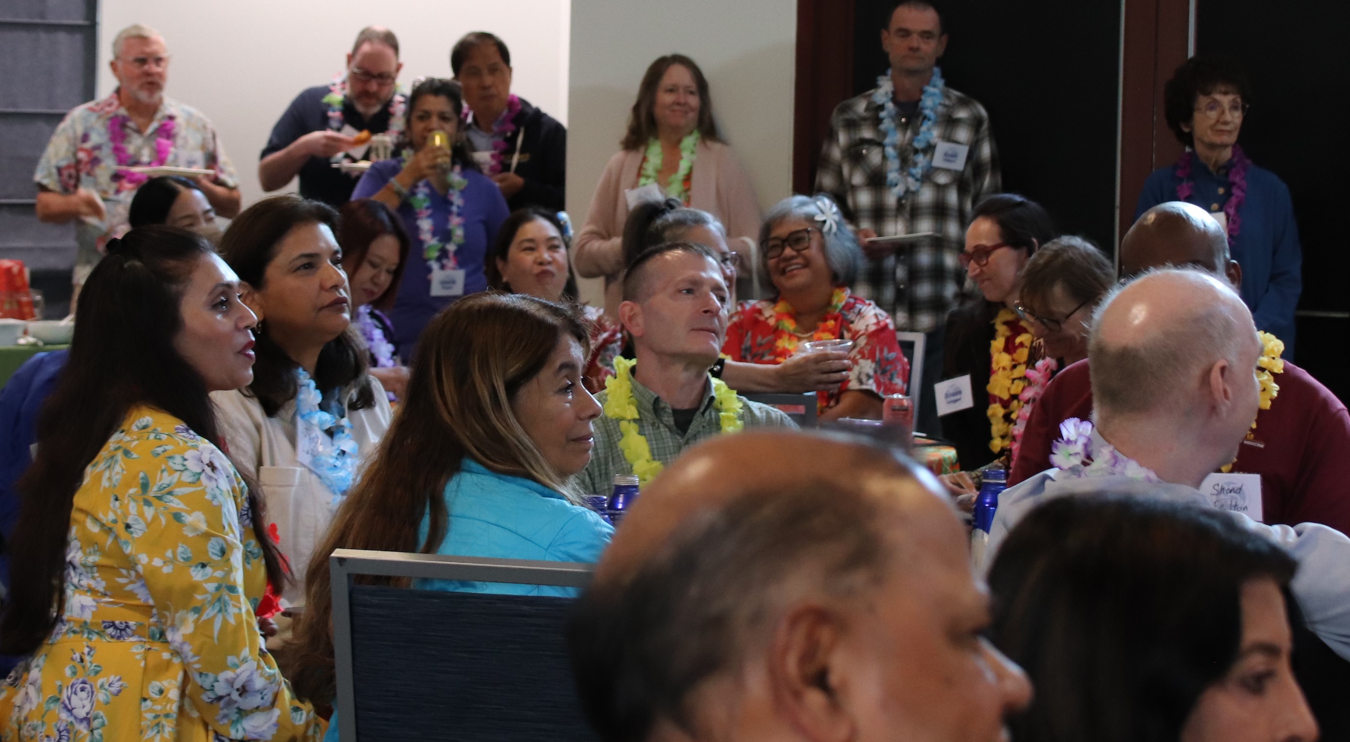 People wearing luau-themed outfits sit at tables and listen to someone speak off camera.
