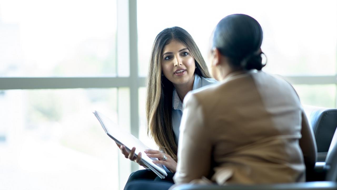 Photo of person holding a clipboard and focused intently on someone, who has their back turned to the camera. 