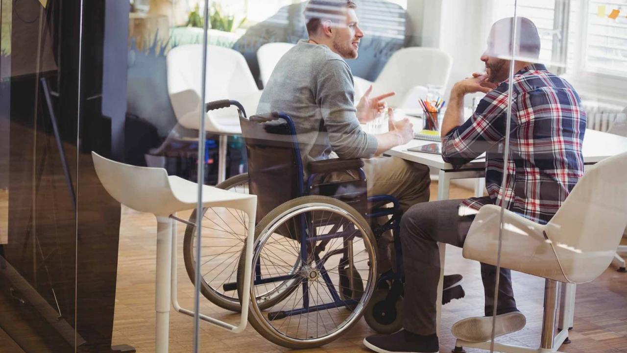 Person in wheelchair at a desk talking to someone else.