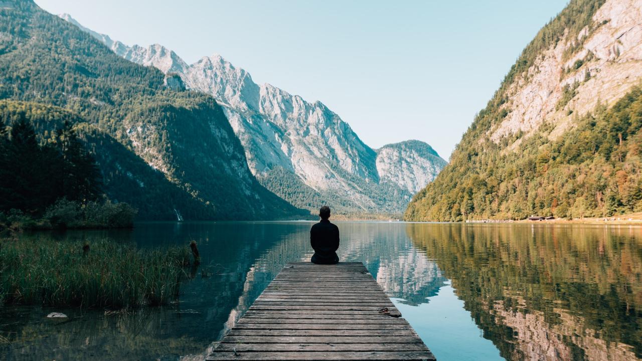 Person sitting on a wooden dock by a tranquil lake, surrounded by mountains and greenery.
