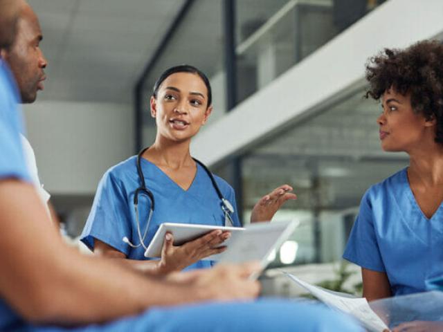 Female and male nurses wearing blue scrubs hold tablets and papers while talking.