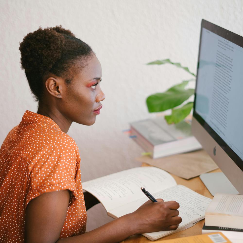 A woman sits at her desk writing in a notebook. She is looking at a computer screen in front of her.