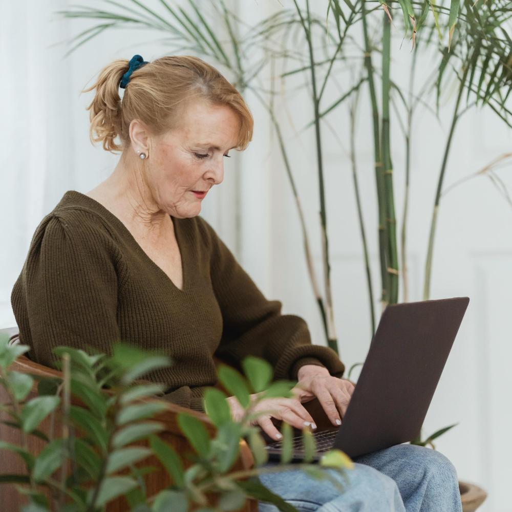 An older woman sits in an armchair while using a laptop in her lap.