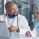 A male doctor talks to a female colleague wearing a hijab, both engaged in conversation by a laptop.