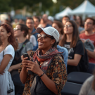 Outdoor crowd at evening event, smiling woman in white cap holding phone