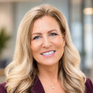 Professional headshot photograph of a smiling middle-aged woman with long wavy blonde hair, maroon blazer
