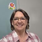 Smiling woman with glasses in a plaid shirt, colorful flower decoration in the background.