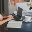 Hands typing on a laptop on a black table. Also on the table are a teacup, an open notebook, and a flower vase with white roses in it.