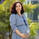 Photo of Caroline wearing a blue dress and a UC Davis Health badge, smiling at camera with hands loosely clasped at their stomach. 