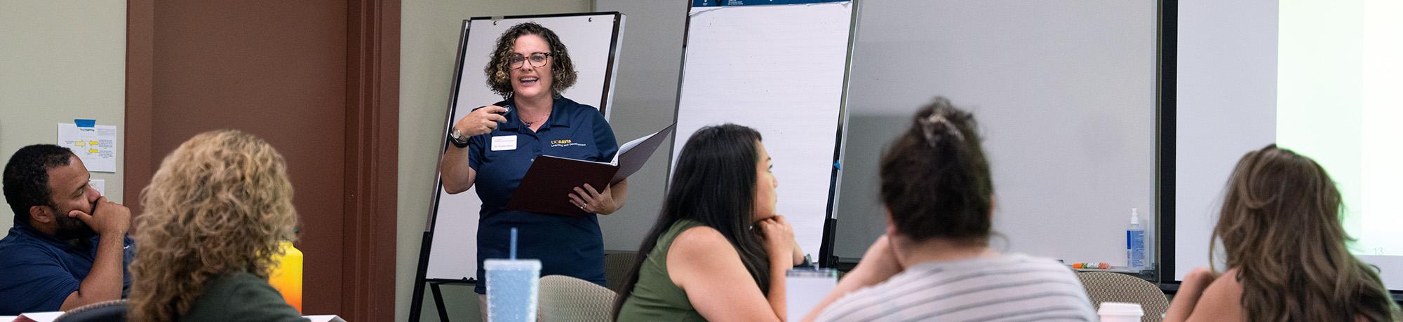A woman stands at the front of the room with a folder and presents to a group of people.